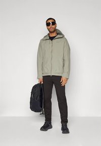 Bărbat îmbrăcat cu o jachetă cu glugă de culoare gri deschis, pantaloni negri, adidași negri și ochelari de soare mari, ținând un rucsac negru într-o mână.