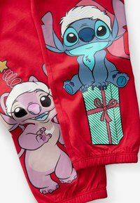 Pantaloni sportivi rossi con personaggi dei cartoni animati Stitch e Angel, con colori vivaci e motivi festivi, tra cui un design a scatola regalo verde.
