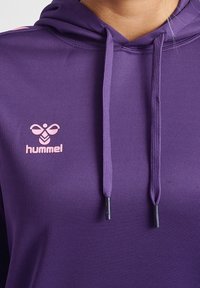 Detailní záběr na fialovou mikinu s kapucí a růžovým logem Hummel ve tvaru včely na hrudi.