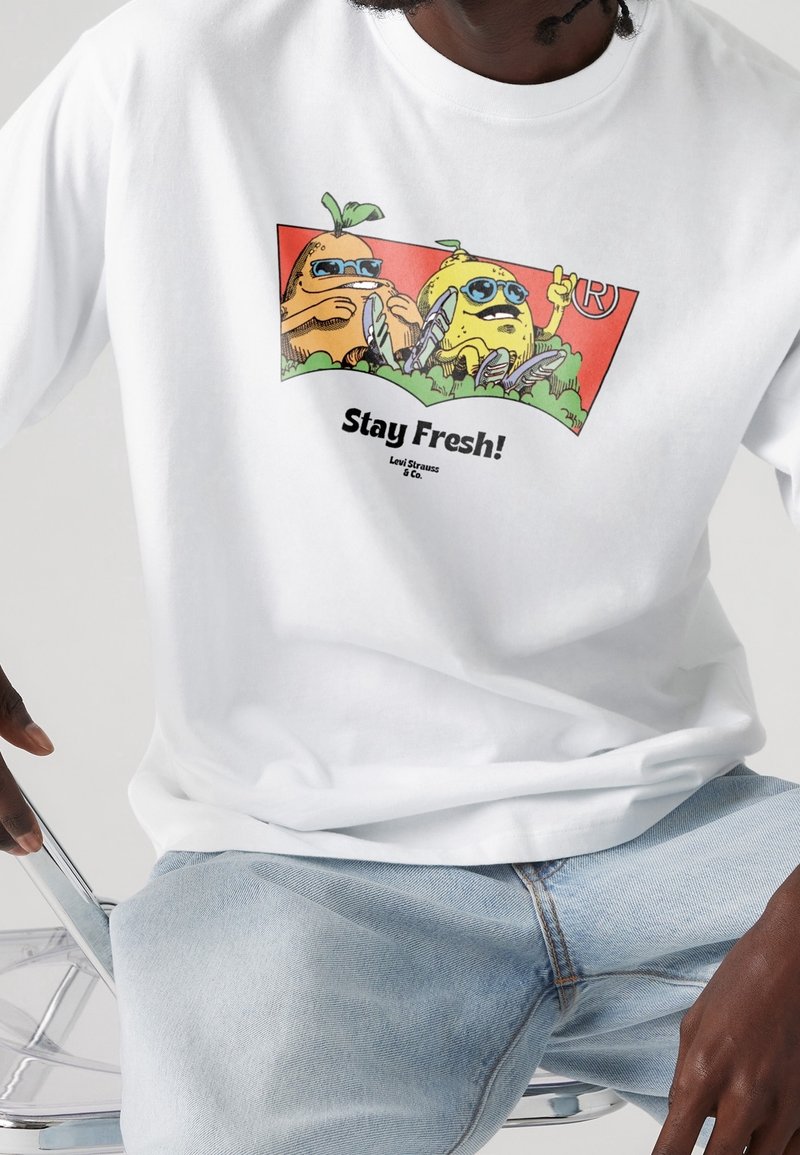 Deux fruits dessinés portant des lunettes de soleil et des baskets avec le texte « Stay Fresh ! Levi Strauss & Co. » sur un T-shirt blanc et un jean bleu clair.