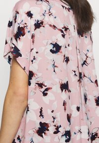 Femme portant un tissu rose ample avec un grand motif floral blanc et bleu foncé, montrant une partie de son bras et ses cheveux bruns.