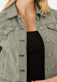 Olivgrön denimjacka med krage, silverfärgade tryckknappar, två bröstfickor och en figurnära design. Materialet har en mjuk textur.