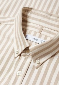 Camisa a rayas beige y crema con cuello tipo solapa, que presenta detalles de botones y una tela texturizada. Etiqueta visible con las iniciales de la marca.
