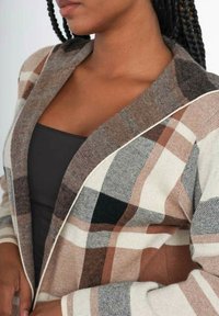 Blazer à carreaux avec des tons beige, marron et noir, en tissu texturé, col châle et finition blanche le long des bords.