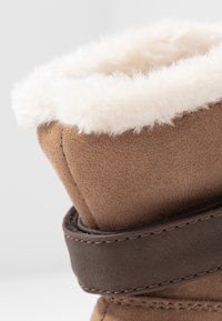 Brązowe zamszowe botki sięgające kostki z białym sztucznym futerkiem na górze i ciemnobrązowym skórzanym paskiem wokół kostki, z widocznymi szwami.