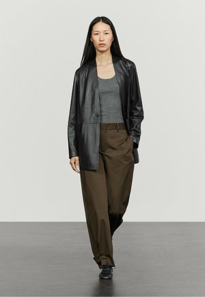 Femme aux longs cheveux foncés portant une veste en cuir noire, un débardeur gris, un pantalon marron ample et des chaussures noires, marchant sur un sol gris.
