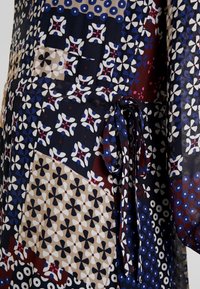 Gros plan sur un tissu à motifs floraux géométriques en bleu marine, beige, blanc, rouge et bleu, avec une boucle de ceinture en tissu nouée sur le côté.