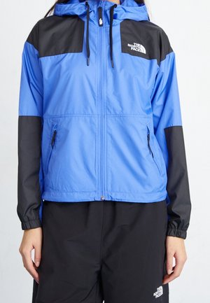 Blå og sort North Face jakke med frontlynlås, hætte med snore, lynlåslommer i siderne, båret med sorte bukser.