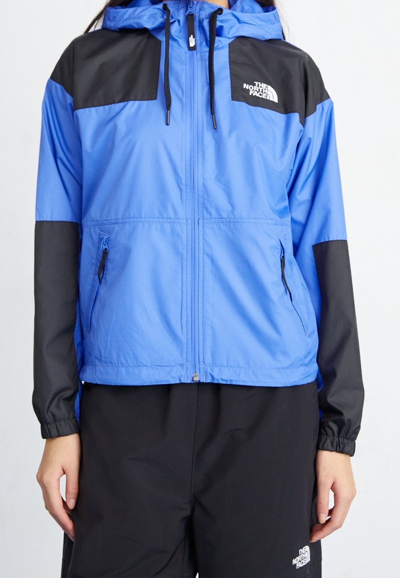 Veste North Face bleue et noire avec fermeture éclair à l'avant, capuche avec cordons, poches latérales zippées, portée avec un pantalon noir.