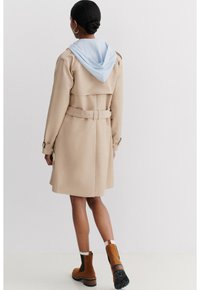 Beige Trenchcoat mit einer hellblauen Kapuze, tailliert mit einem Gürtel und geknöpften Manschetten. Kombiniert mit braunen und schwarzen Stiefeletten. Glatte Textur.