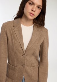 Blazer aderente marrone realizzato in tessuto morbido, con rever a scollatura, chiusura frontale a due bottoni e due tasche laterali. Texture liscia con dettagli sottili.