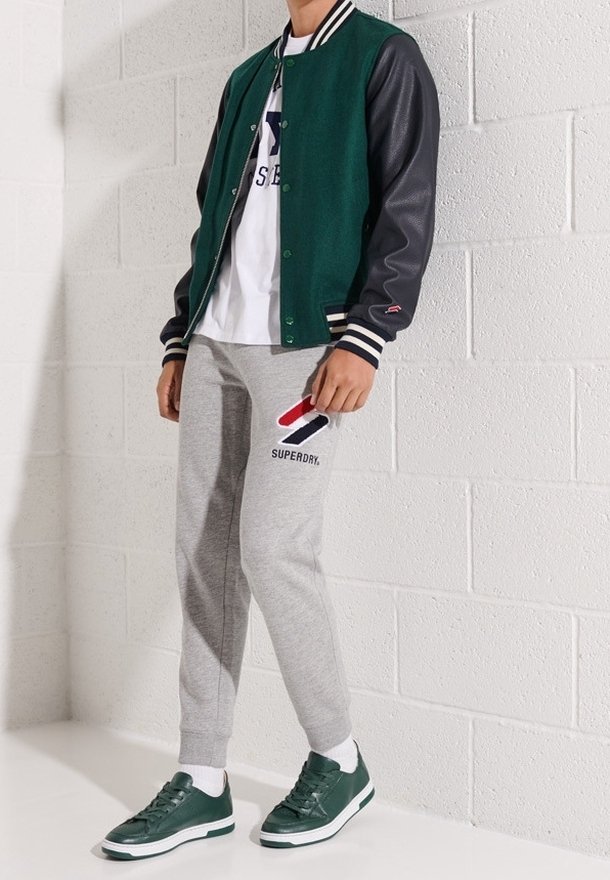 Personne portant une veste varsity verte et noire, un t-shirt blanc, un pantalon de survêtement gris Superdry et des baskets vertes, debout contre un mur en briques blanches.