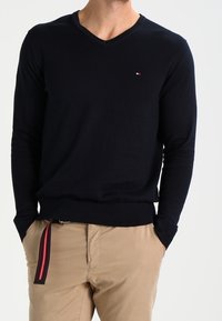 Pull col en V marine en maille fine, manches longues et petit logo de marque sur la poitrine. Porté avec un pantalon beige et une ceinture rayée.