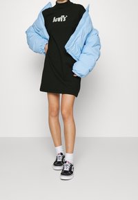 Personne portant une robe noire Levi's, une doudoune bleu clair posée sur les épaules, des chaussettes blanches et des baskets Vans noires sur un fond uni.