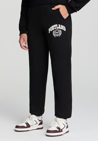 Czarne dresy z elastycznym pasa i bocznymi kieszeniami, z białą grafiką "PORTLAND" oraz motywem laurowym z przodu.