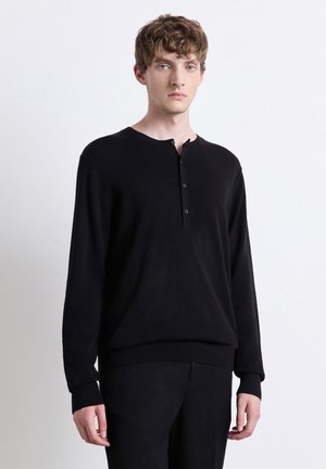 Junger Mann mit heller Haut und lockigem Haar, der ein schwarzes Langarm-Henley-Shirt und schwarze Hose trägt, steht vor einem schlichten weißen Hintergrund.