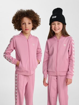 Zwei Kinder in passenden rosa Trainingsanzügen mit weißen Chevron-Streifen, eines mit lockigem Haar, das andere trägt eine schwarze Strickmütze, lächelnd.