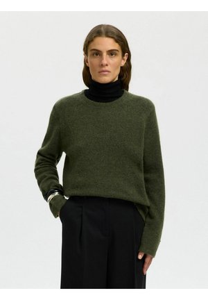 Femme portant un pull vert foncé par-dessus un col roulé noir, avec un pantalon noir, debout avec une main dans la poche sur un fond clair.
