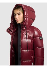 Vínová péřová bunda s lesklým prošívaným povrchem, velkou kapucí, zapínáním na zip a černými kontrastními detaily. Na rukávu je nášivka s logem.