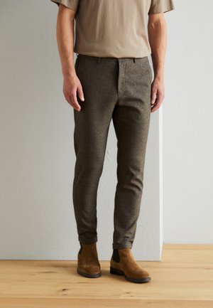 Uomo con pantaloni marroni slim-fit, t-shirt beige e stivali Chelsea in camoscio color nocciola, in piedi su pavimento in legno contro parete bianca.