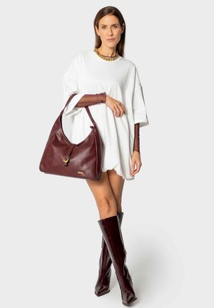 Vrouw in oversized witte jurk met bruine mesh mouwen, bruine laarzen tot de knie, gouden ketting, en houdt een grote bruine leren handtas vast.