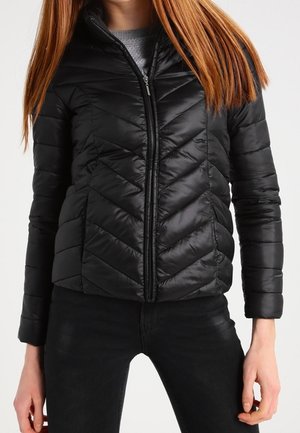 Woman wearing a black quilted zip-up jacket with chevron stitching and black pants, standing with hands relaxed at sides.