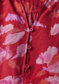 Rote Bluse mit floralen Mustern in Rosa und Lila, V-Ausschnitt und passenden Knöpfen entlang der Vorderseite, aus leichtem Stoff gefertigt.