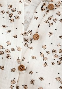 Lätt texturerad tygskjorta i krämfärg med små beige blommönster, med bruna knappar fram och krage.