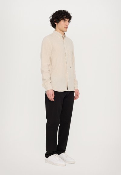 Chemise en lin beige à boutons avec une poche poitrine unique, associée à un pantalon noir et des baskets blanches, présentée sur une silhouette debout.