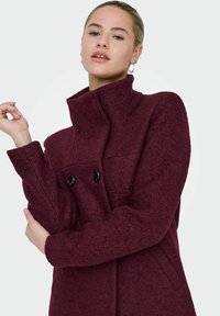 Burgundijas vilnas sajaukuma mēteli ar augstu apkakli, divām melnām pogām un teksturētu audumu. Garas piedurknes un sānu kabatas.