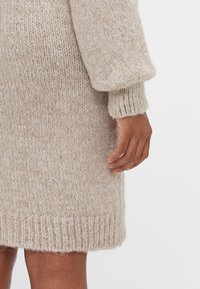 Bež ple knitted kleit, millel on tekstureeritud viimistlus, ribikätikud ja äär. Disainil on katkematu lõige ja pikad, volüümsed varrukad.