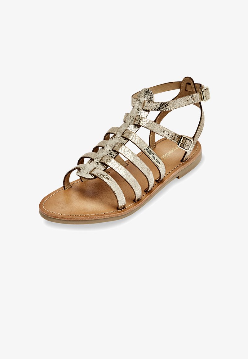 Sandale dorée à lanières avec une semelle plate, présentant six bandes métalliques et des brides à la cheville avec des boucles, sur une semelle en cuir beige.