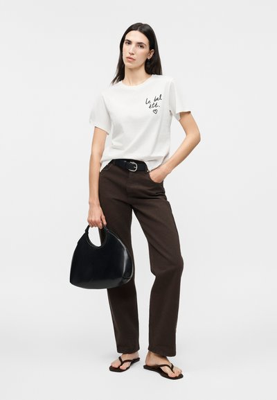 Γυναίκα που φορά λευκό t-shirt με μαύρο κείμενο, σκούρο παντελόνι, μαύρη ζώνη, μαύρα σαγιονάρες, κρατώντας μαύρη τσάντα σε μονόχρωμο φόντο.