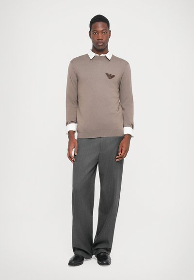 Brown knit sweater with ribbed hem and seven sleeves, featuring a dark logo on the chest. Paired with gray wide-leg trousers and black shoes.
