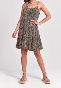 Frau trägt ein ärmelloses, leopardgemustertes, knielanges Kleid mit beigen, geflochtenen Plateausandalen und steht mit einer Hand auf der Hüfte.