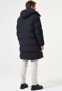 Abrigo acolchado de un oscuro color navy con capucha, con un diseño acolchado y puños elásticos. Combinado con pantalones en un tono beige claro y calzado negro.