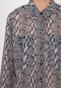 Chemise boutonnée avec motif peau de serpent gris et noir, dotée de deux poches à rabat à l'avant et d'un col classique.