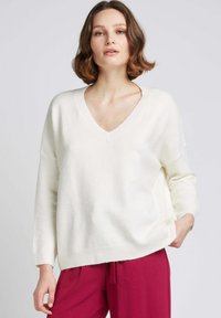 Pull blanc à col en V avec une coupe ample, des manches longues et un ourlet côtelé, associé à un pantalon fuchsia. Texture douce, détails minimaux.