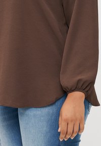 Gros plan sur la main d'une personne tenant l'ourlet d'une chemise marron à manches longues, associée à un jean bleu. Le tissu semble texturé.