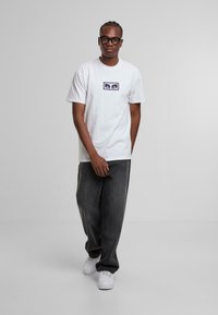 Hvid t-shirt med indrammet grafisk design på brystet, parret med løse grå denimbukser og hvide sneakers. Enkelt, afslappet stil.
