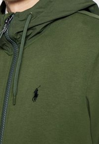 Grön zip-huvtröja tillverkad av ett mjukt material, med en snörning i huvan och en liten svart logotyp på bröstet.