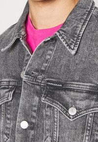 Giacca di denim grigia con colletto a punta, due tasche frontali e chiusure con bottoni in argento. Indossata sopra una maglietta rosa brillante. Etichetta del logo visibile.