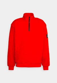 Punainen fleece-poolopaita korkealla kauluksella ja puolivetoketjulla. Ominaisuuksiin kuuluvat resorireunukset hihoissa ja vasemmalla puolella oleva brändätty kangasmerkki. Pehmeä rakenne.