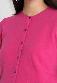 Ljusrosa cardigan gjord av mjukt tyg, med en rund halsringning och sju knappar framtill för stängning.