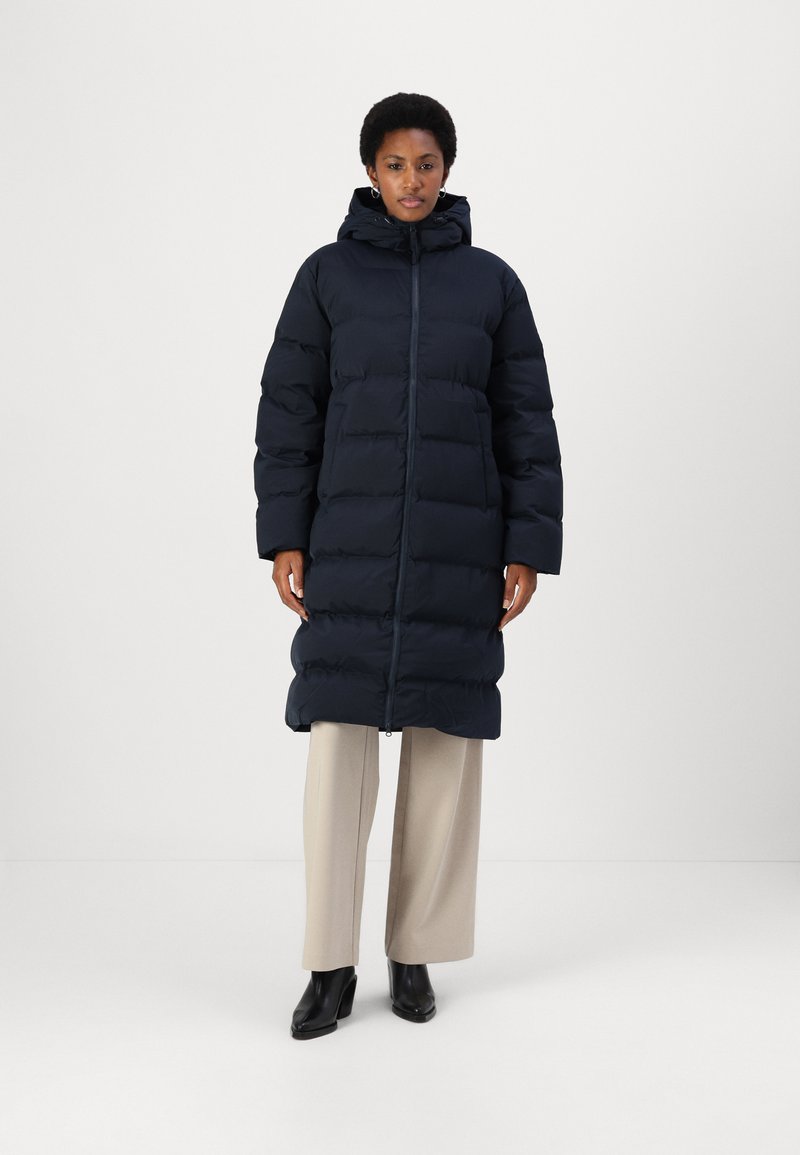 Navy puffer coat with a hood, featuring a quilted design and full-length zipper. Worn over cream wide-legged trousers and black boots.