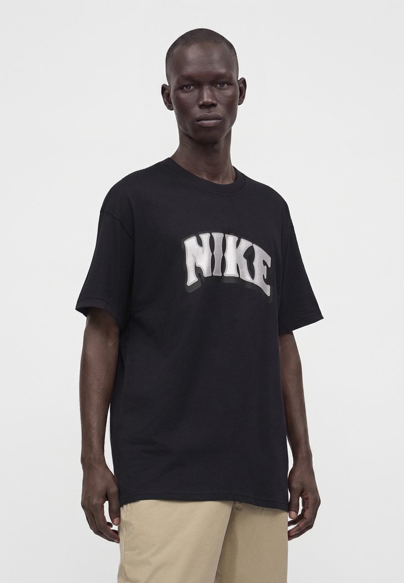 Uomo che indossa una maglietta nera oversize Nike e pantaloni beige, in piedi davanti a uno sfondo chiaro e semplice.