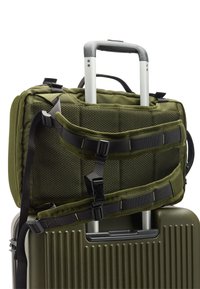 Zielona oliwkowa torba podróżna z siateczkowym panelem, regulowanymi paskami i zapięciem na klamrę, umieszczona nad teksturowaną, ciemnozieloną walizką twardą.