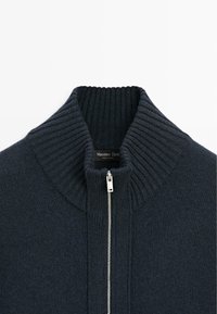 Maglione a coste grigio scuro con colletto alto e zip argento, caratterizzato da una texture morbida e un design minimalista.