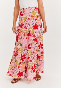 Žena v ružovej maxi sukni s kvetinovým vzorom obsahujúcim červené, broskyňové a zelené kvety, skombinovanej s hnedými sandálmi a bielym tričkom.