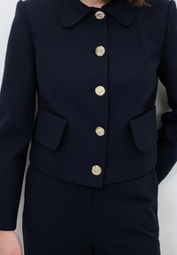 Marinblå skräddarsydd kavaj med rundad krage, sex guldfärgade texturerade knappar och två framfickor. Slätt tyg med en strukturerad design.